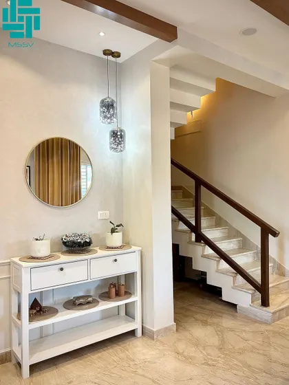 The entryway sets a welcoming tone with a white console table, a gold-framed mirror, and elegant hanging lamps. The marble staircase with a wooden railing adds a touch of classic sophistication.