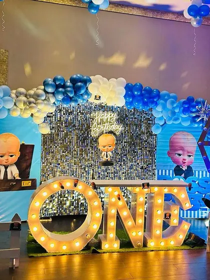 A Boss Baby first birthday party with a silver shimmer wall, "ONE" marquee letters, and a white picket fence.
