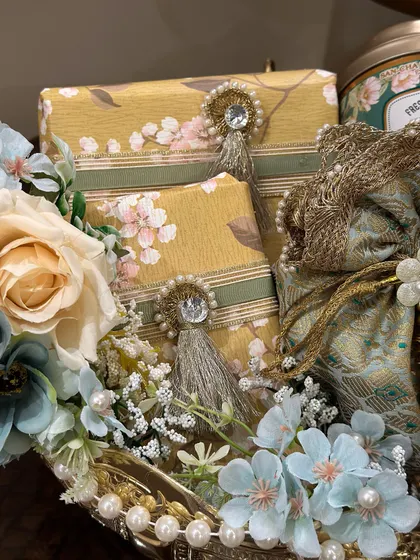 A close-up view of the details in our Diwali hamper. The combination of floral-wrapped gifts with jewel tassels, a brocade potli, and a mix of blue and cream flowers creates a rich texture.