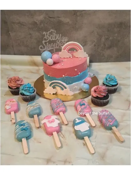 A full dessert spread for a baby shower. This set includes a rainbow themed cake, matching cupcakes, and adorable pink and blue cakesicles decorated with baby clothes and bottle shapes.
