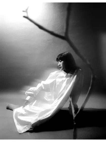 A black and white film photograph where the model sits on the floor, with the shadow of a branch creating a stark graphic element. The composition is minimalist and evocative, telling a story with light and shadow.