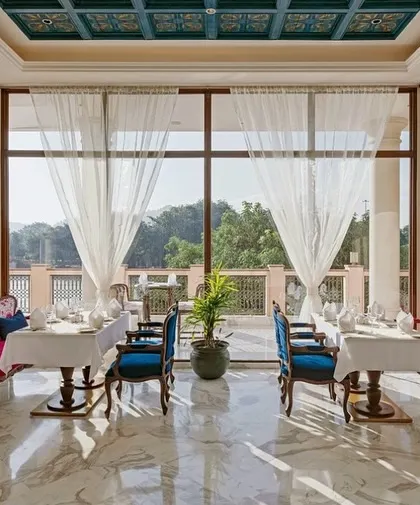A sun-drenched dining area at IHCL Pilibhit-Haridwar, where floor-to-ceiling windows frame the lush outdoors. The vibrant pink and blue seating adds a pop of color to the serene, marble-floored space.