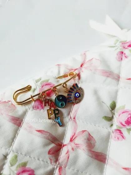 Pin your vibe. These safety pin charms are fully customizable. This one features 'HAPPY' letter charms, a yin-yang, and an evil eye for a mix of fun and protection.