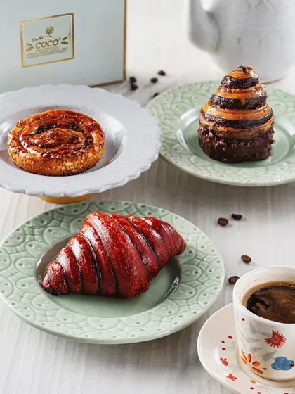 A delightful spread of pastries from The Coco Company, including a red croissant and a chocolate swirl. Paired with a cup of black coffee, this bright and airy shot creates a perfect morning coffee-and-pastry scene.