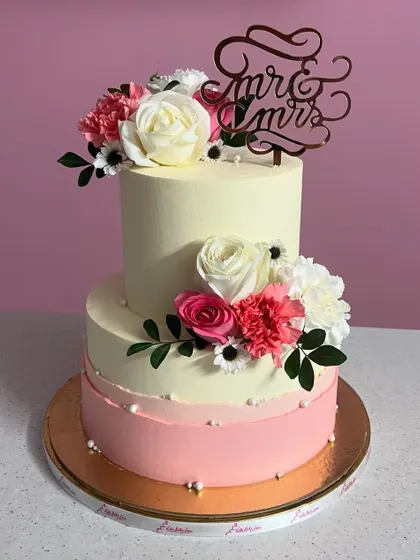 A lovely two-tiered anniversary cake with a pink ombre effect. It's decorated with fresh white and pink flowers and a "Mr & Mrs" topper, celebrating love in the sweetest way.