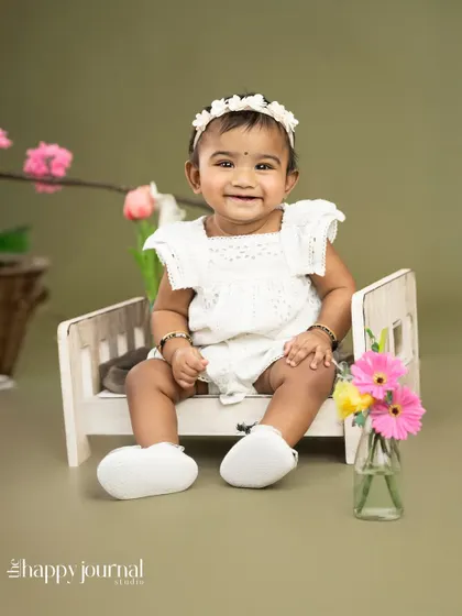 A smiling baby girl in a white outfit, sitting on a miniature wooden bed. The floral props add a touch of spring to this sweet milestone photo.