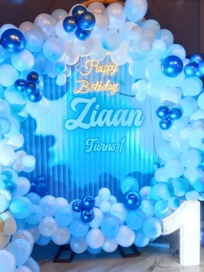 A close-up of a blue-themed first birthday for Ziaan. The backdrop features a cloud-like design, complemented by a full arch of blue and white balloons and a cute teddy bear.