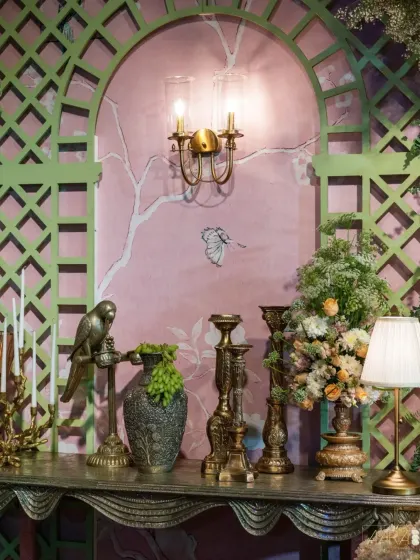 A beautiful vignette from the 'Whispers of Spring' event. A console table is styled with antique brass candelabras, a parrot figurine, and lush florals, all set against a sage green trellis and a hand-painted wall.