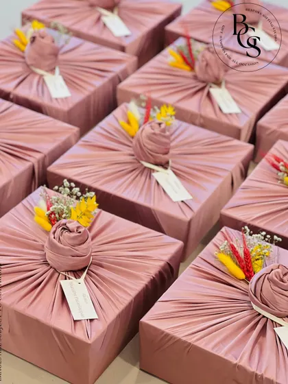 A batch of gifts elegantly wrapped in dusty rose fabric. Each gift is topped with a fabric rose knot and a small sprig of colorful dried flowers, creating a cohesive and beautiful presentation for a special event.