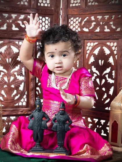 A one-year-old girl celebrating her milestone with a Vitthal theme. Dressed in a bright pink paithani-style dress, she blesses the camera with a wave.