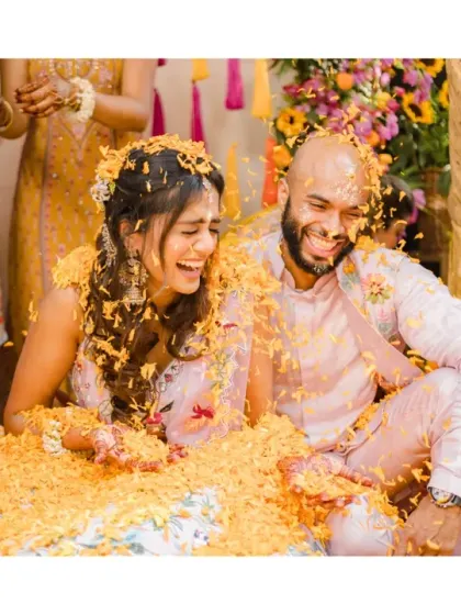 The couple laughs uncontrollably as they are buried in a mountain of marigold petals. This image is the definition of "phoolon ki haldi" and pure, uninhibited joy.