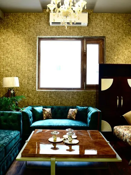 A formal living room designed with a rich, jewel-toned palette. The space features a custom L-shaped sofa in deep green velvet, a solid wood coffee table, and floral accent chairs, all set against a textured gold wallpaper.