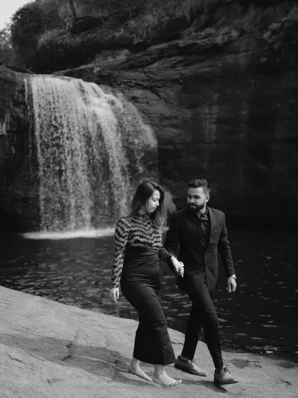 A dramatic black and white image of a couple walking by a waterfall, highlighting the contrast and texture of the natural landscape.