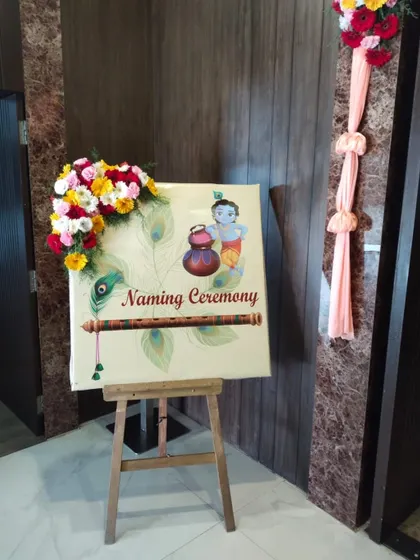 An elegant welcome easel for a naming ceremony, decorated with a fresh flower arrangement. The sign features a traditional Krishna illustration, setting a blessed tone for the event.