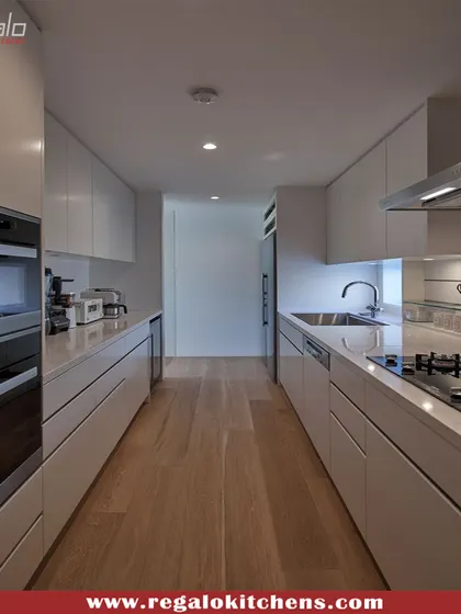 An ultra-minimalist parallel kitchen in all-white. The handle-less design and integrated appliances create a seamless and clean look, making the narrow space feel larger and brighter. The wooden floor adds a touch of warmth.