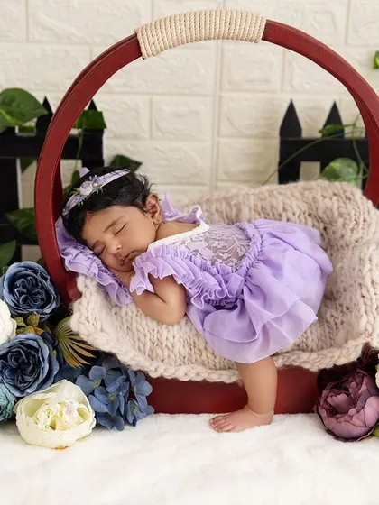 A beautiful newborn girl in a floral setting. Dressed in a delicate lavender outfit, she is peacefully asleep in a basket surrounded by colorful flowers.