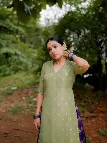 A full-body portrait of actress Anupama Gowda in a traditional outfit. The natural, earthy tones of the location complement her elegant attire.