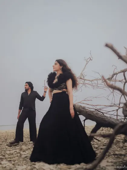 A high-fashion, editorial-style shot on a desolate beach. The couple's dramatic black outfits contrast beautifully with the natural, raw texture of the landscape.