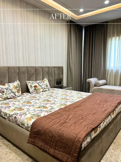 The "after" view of a bedroom renovation, now featuring a calm and soothing design with a plush headboard, neutral tones, and a comfortable chaise lounge by the window.