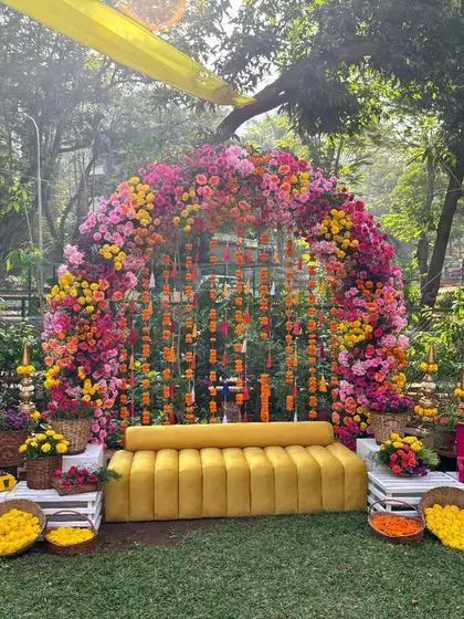 A vibrant and colorful Mehendi seating area, featuring a massive floral arch of pink, yellow, and orange blooms, with a comfortable yellow sofa for the bride.
