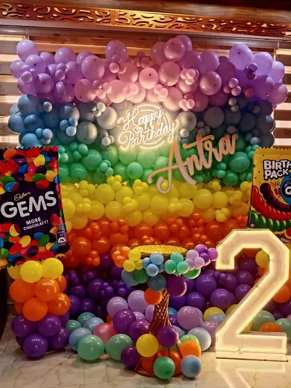 A super colorful and fun Gems theme for a 2nd birthday. This vibrant setup includes a full wall of rainbow balloons, a neon sign, a light-up number, and giant Gems candy packet props.