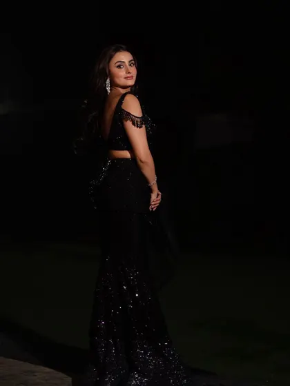 A classic over-the-shoulder pose that highlights the backless design and tassel details of the saree blouse. The lighting carves the model out of the darkness.