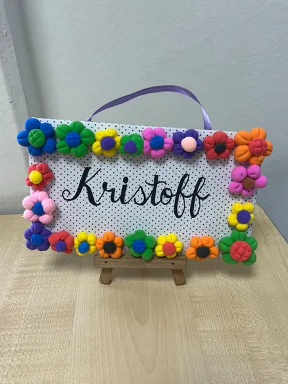 This personalized nameplate is decorated with colourful flowers made from clay. It's a fun project that helps kids with their fine motor skills and creativity.