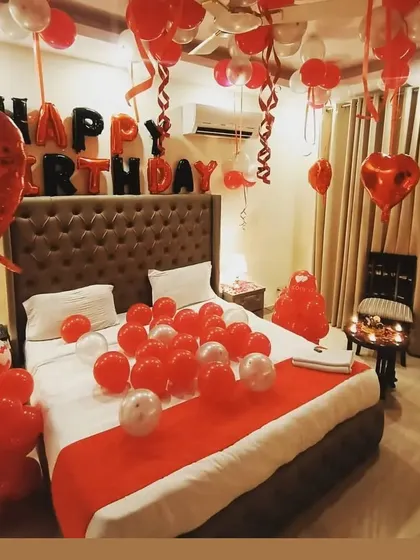 A hotel room decorated for a birthday surprise, with red and white balloons, "Happy Birthday" foil letters, and a neatly decorated bed.