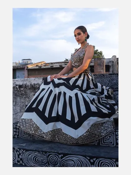 A seated portrait of a model in a Vandana Sethi lehenga, showcasing the full volume and pattern of the skirt. The rooftop setting provides a unique, urban backdrop for the ethnic design.
