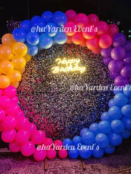 A vibrant, multi-color balloon ring set against a silver sequin wall. The 'Happy Birthday' neon sign in the center makes this a fun and eye-catching photo spot.