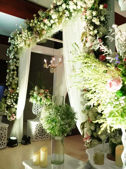 A close-up of a romantic floral arch, made with white flowers and baby's breath. A small chandelier hangs within the arch, adding a touch of classic elegance to the design.