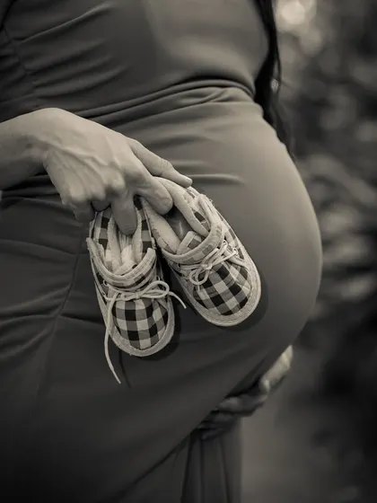 A close-up, black and white shot focusing on a pair of tiny plaid baby shoes held against the baby bump. A classic and artistic detail shot.