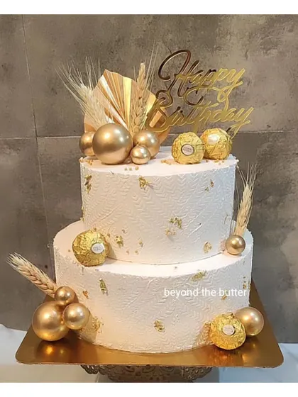 A luxurious two-tier cake featuring a textured white finish and gold accents. It's decorated with Ferrero Rocher chocolates, gold spheres, and dried wheat for a modern and celebratory look.