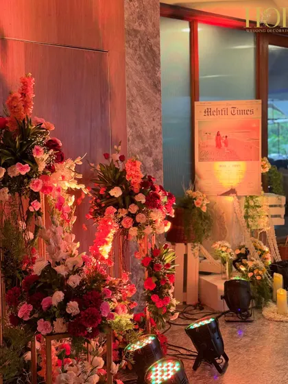 A rustic and charming welcome setup featuring a personalized "Mehfil Times" newspaper sign, surrounded by floral arrangements in wicker baskets and warm candle lights.