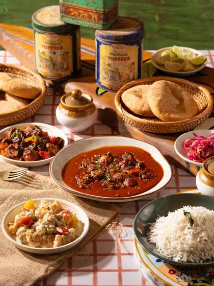 A spread of Goan food, including a rich curry, rice, and bread. The checkered tablecloth gives it a homey, rustic cafe vibe.