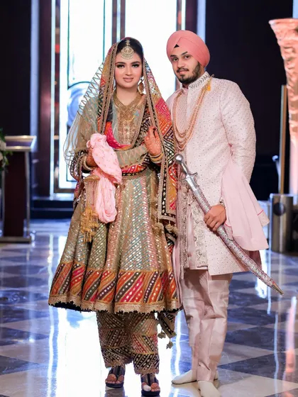 A full-length portrait of the Sikh couple in their unique, colorful wedding attire. The groom holds the kirpan, and the bride's outfit is a beautiful tapestry of colors.