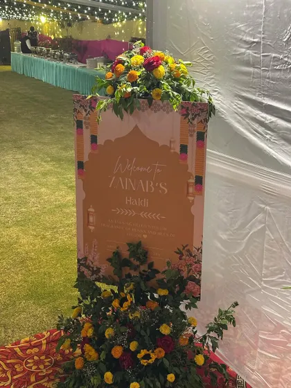 A personalized welcome sign for 'Zainab's Haldi'. The sign is decorated with a beautiful floral arrangement, setting a warm and inviting tone for the event.