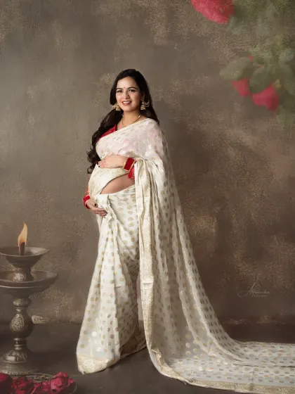 A serene and beautiful solo portrait. The mom-to-be is holding a traditional lamp (diya), adding a spiritual and peaceful element to her white and red saree look.