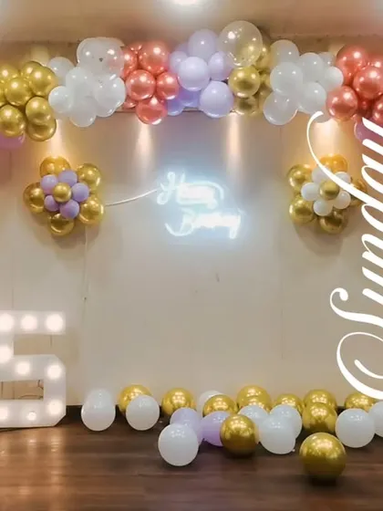 A grand setup for a 75th birthday in a hall, with a large pastel and gold balloon arch, a neon sign, and big light-up marquee numbers.