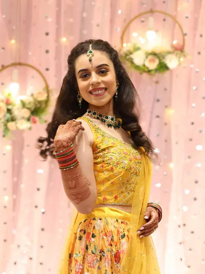 The bride looking radiant in her yellow floral lehenga for the sangeet. The makeup is fresh and bright, with a pop of color from the green jewelry.