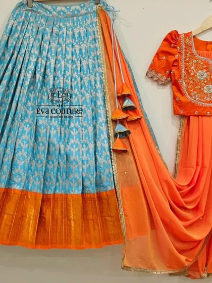 A bright and contrasting pattu lehenga set. The sky blue Banarasi skirt is paired with a vibrant orange blouse with maggam work and an attached orange dupatta.