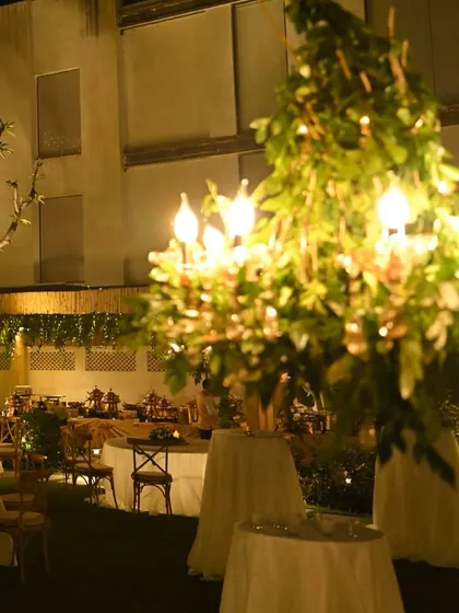 A view of the outdoor party space, with the greenery-wrapped chandelier in the foreground. The composition shows the layers of decor and lighting that create the overall ambiance.