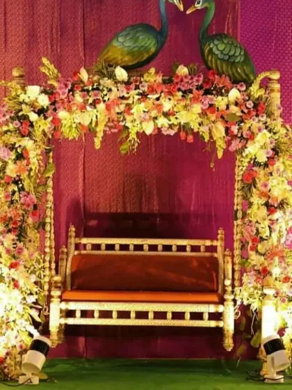 A traditional wooden jhula decorated with a mix of fresh flowers and two peacock figures on top. This kind of setup is perfect for a Mehandi, Sangeet, or even a Janmashtami celebration at home.