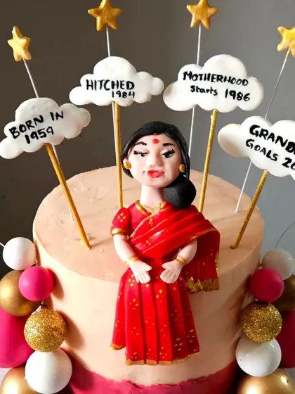 A detailed view of the figurine and milestone markers for the 65th birthday cake. Each cloud is hand-lettered to celebrate a different chapter of her life, from "Hitched 1984" to "Grandma Goals 2022."