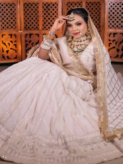 A full-length shot of a bride seated in a stunning all-white lehenga. The pose is regal and serene, set against a backdrop that resembles ancient palace architecture.