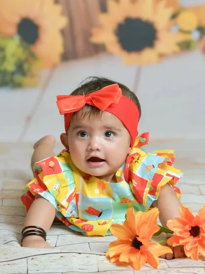 A colorful and happy baby girl in a bright outfit and red bow lies on her tummy during a fun studio session, looking up with an adorable and curious expression.