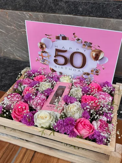 A beautiful floral crate celebrating a 50th birthday. The mix of pink, purple, and white flowers creates a vibrant and joyful arrangement, with a chocolate bar nestled in the center.