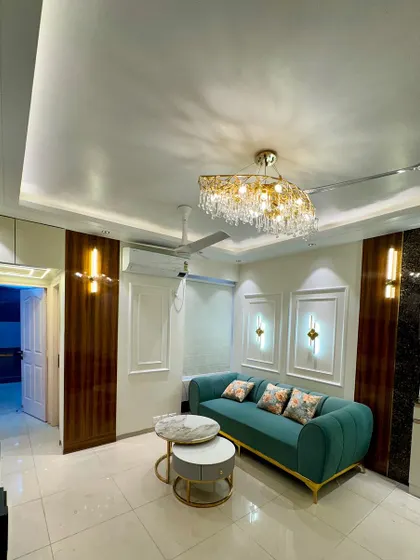 A full view of a luxurious drawing room. The design includes elegant wall moulding, modern sconce lights, and a beautiful crystal chandelier, all complementing the teal sofa.