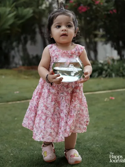 A toddler in a pink floral dress stands in the garden holding a fishbowl. A moment of quiet curiosity and wonder.