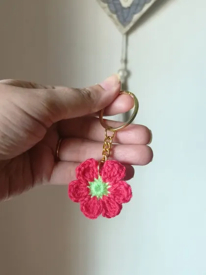 A bright pink floral keychain with a green center, held up to show the detailed crochet work.